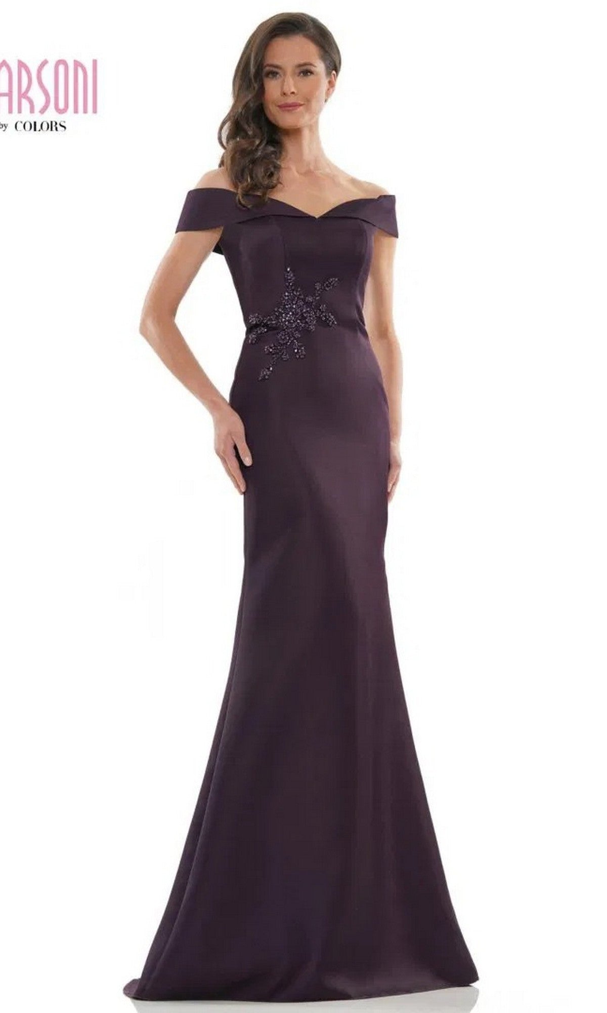 A woman wears the Marsoni MV1003 long formal dress in eggplant satin, featuring an off-the-shoulder neckline and beaded floral embellishment at the waist, standing against a plain background.